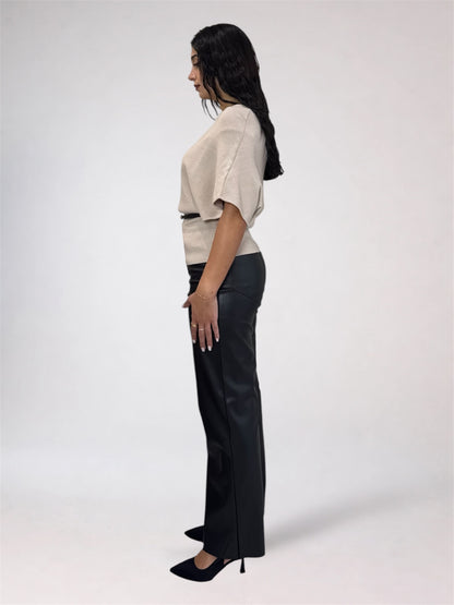 A woman shown in profile wearing a light beige short-sleeve top with a thin dark waistband detail and black wide-leg trousers, standing against a plain light gray studio background.