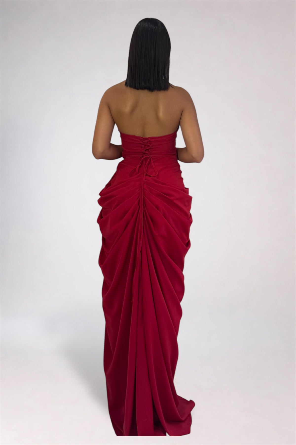 A woman viewed from behind wearing a strapless deep red maxi dress with a gathered ruched back and a long flowing skirt, photographed against a plain light gray studio background.