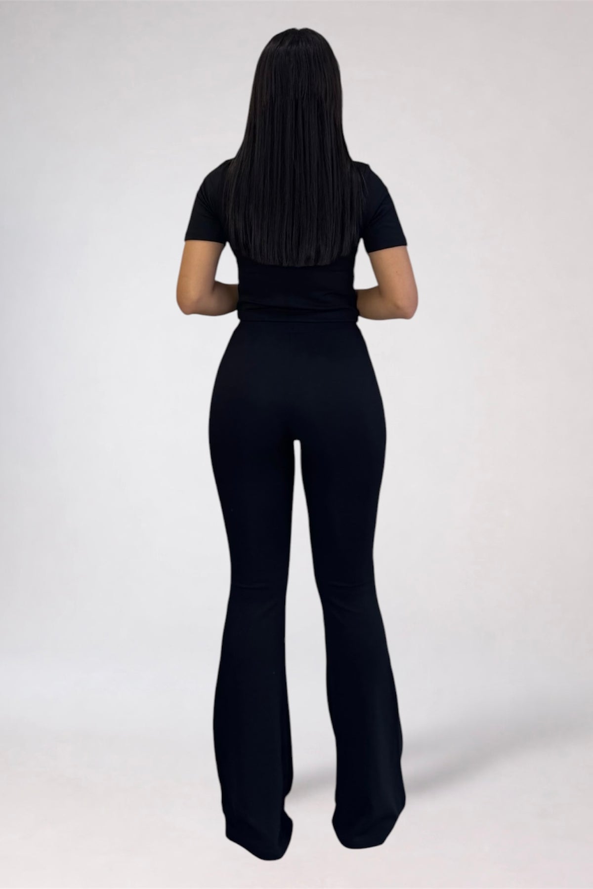A woman viewed from behind wearing a black short-sleeve top and matching black flared pants against a plain light gray studio background.