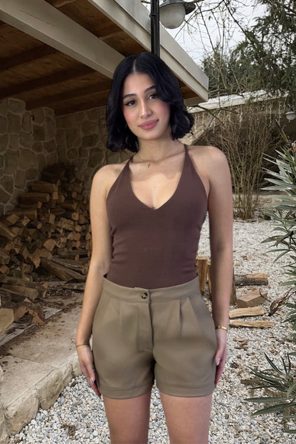 Woman wearing a brown tank top and beige shorts standing outdoors near stacked firewood.