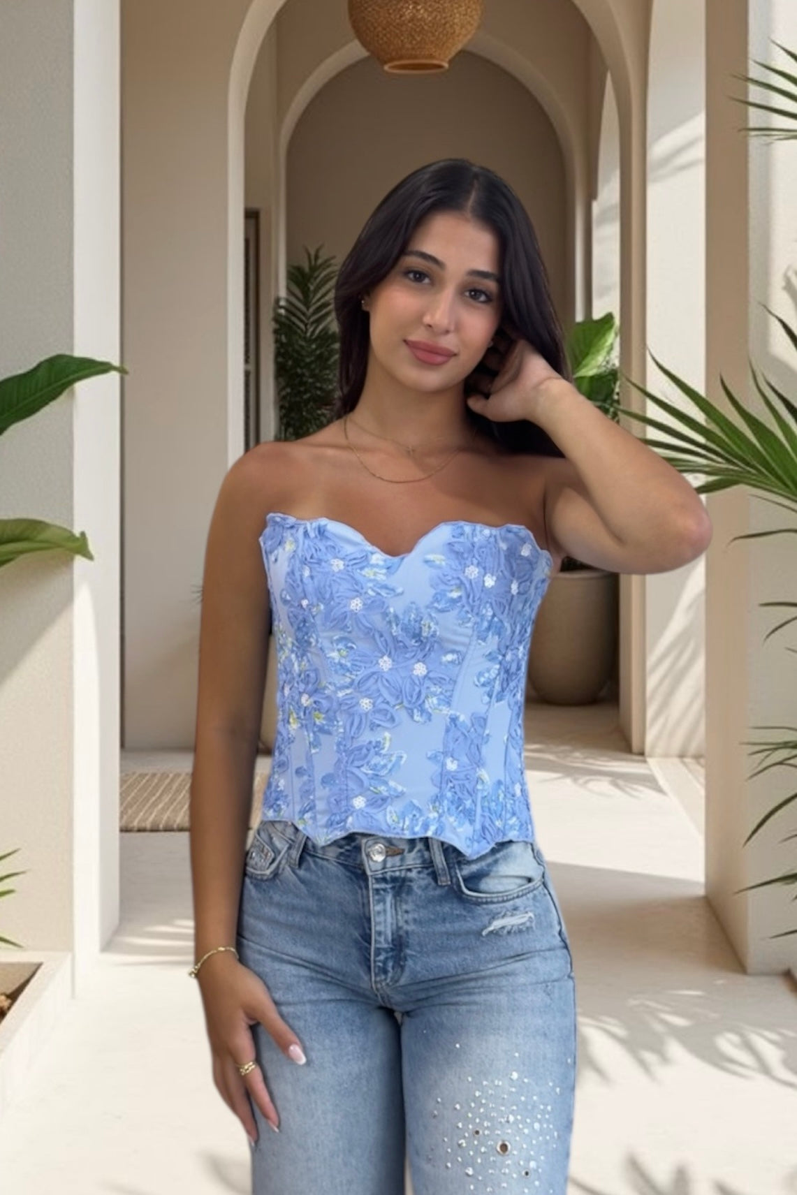 Woman wearing a blue strapless top and jeans standing in a sunlit outdoor setting with plants.