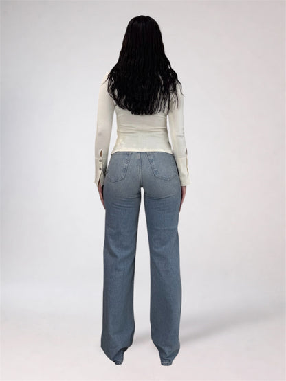 A woman viewed from behind wearing a fitted off-white long-sleeve top and light blue wide-leg jeans against a plain light gray studio background.