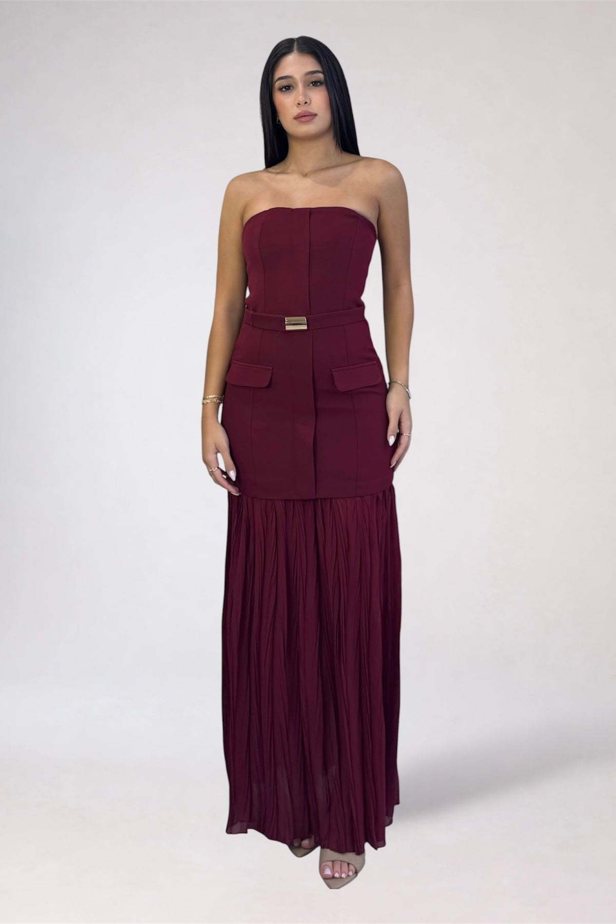 A woman modeling a strapless, deep burgundy maxi dress with a structured corset-style bodice, side pockets, a matching belt with a gold-tone buckle, and a pleated skirt, set against a plain light gray studio background.