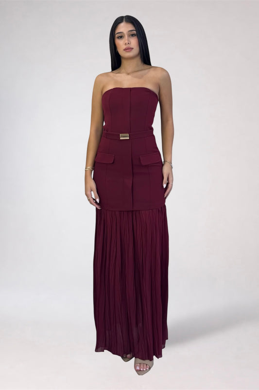 A woman modeling a strapless, deep burgundy maxi dress with a structured corset-style bodice, side pockets, a matching belt with a gold-tone buckle, and a pleated skirt, set against a plain light gray studio background.