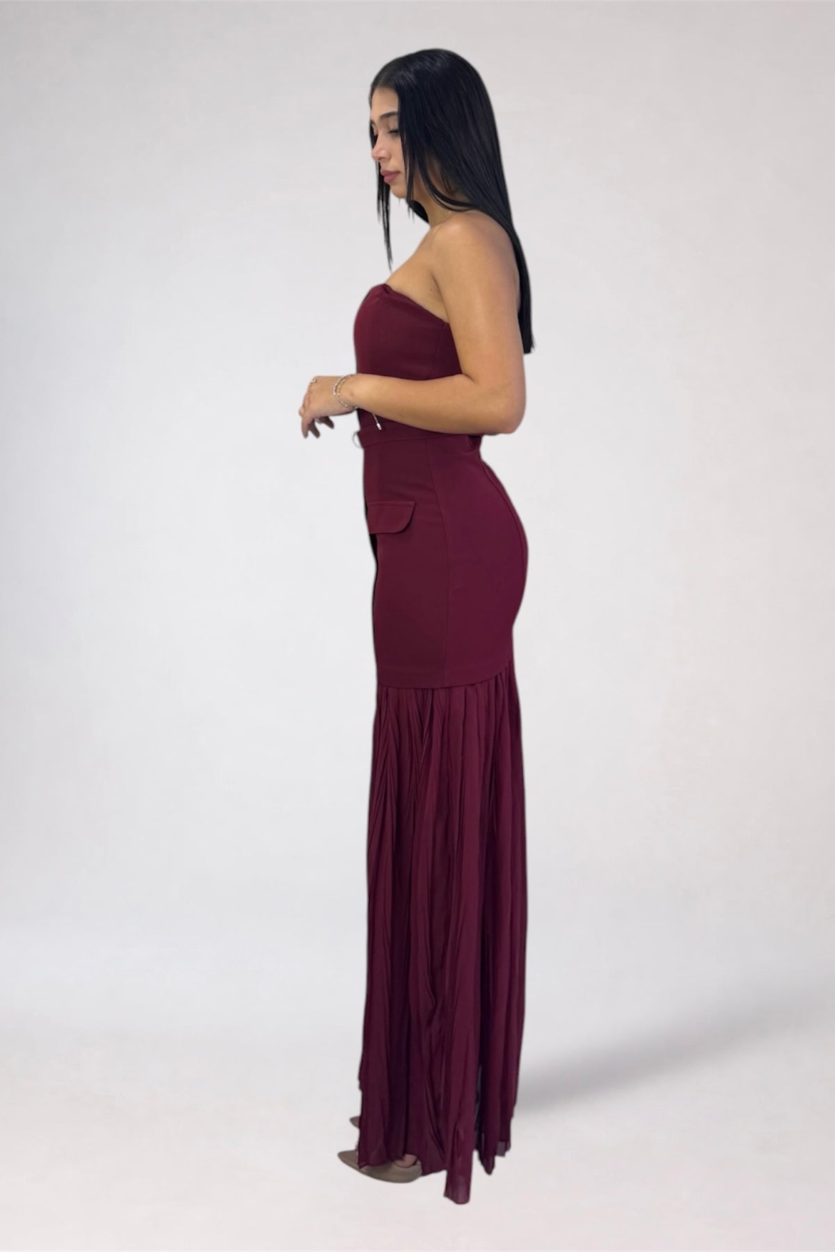 A woman modeling a strapless, deep burgundy maxi dress with a fitted bodice, side pockets, and a flowing pleated skirt, shown in profile against a plain light gray studio background.