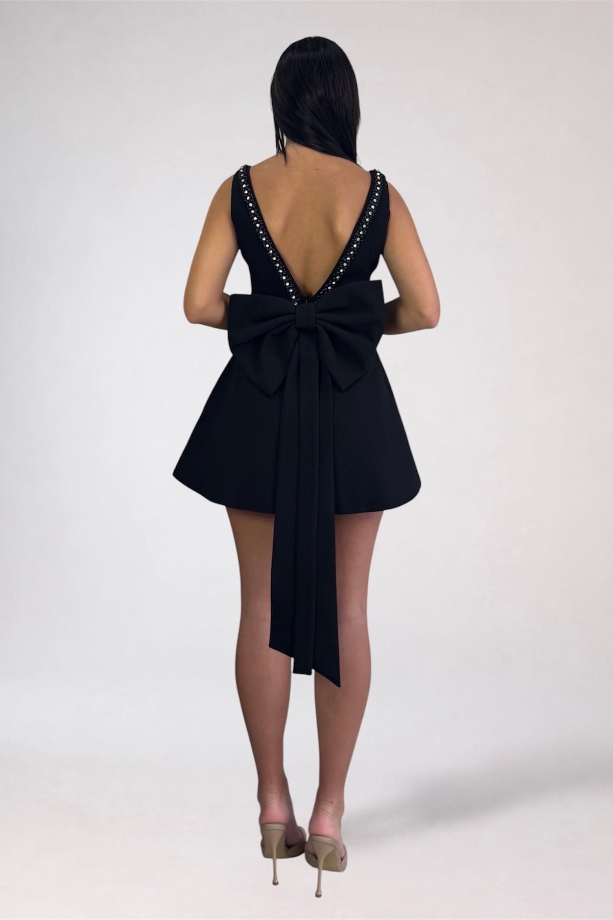 A woman shown from the back wearing a short black dress with a large bow at the back and an open back decorated with small white studs, photographed against a plain light gray studio background.