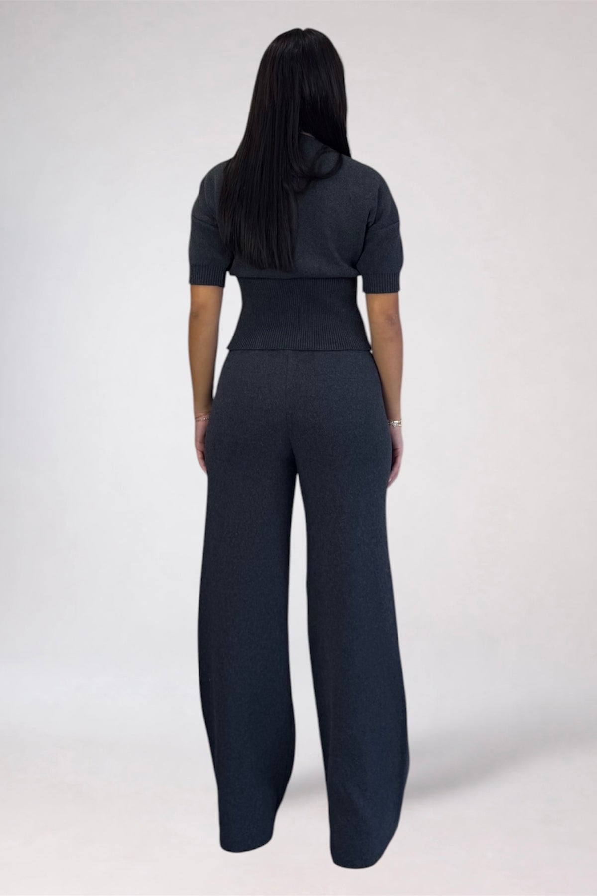 A woman viewed from the back wearing a dark charcoal gray short-sleeve top and matching wide-leg trousers, standing against a light gray studio background.