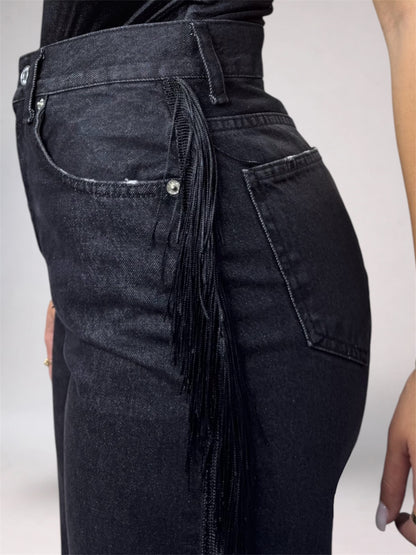Close-up side view of a person wearing dark washed denim jeans with a long fringe detail hanging from the back pocket.
