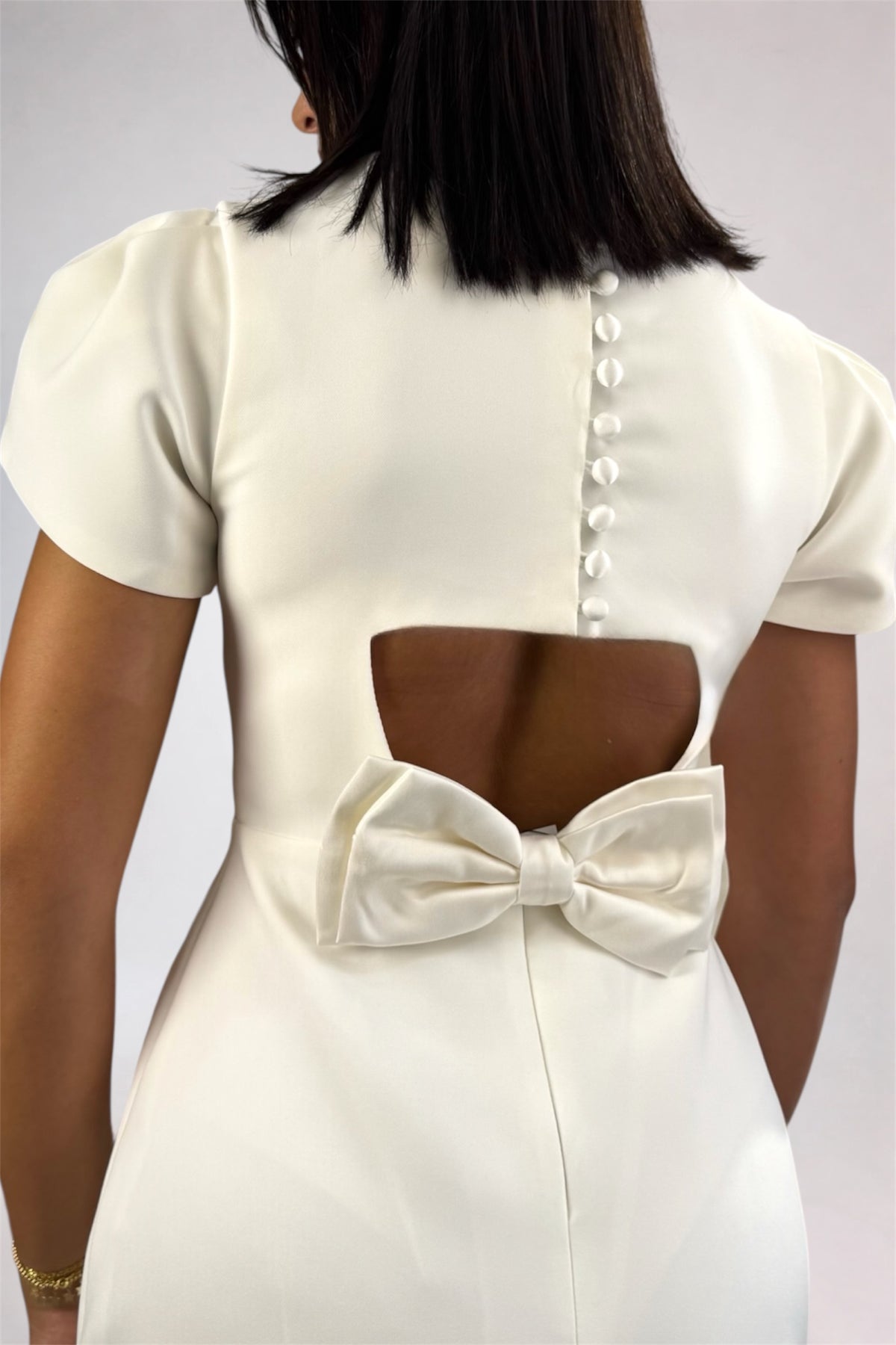 Back view of a woman wearing a cream-colored short-sleeve dress with a large bow and button detail on the back, set against a plain light gray background.