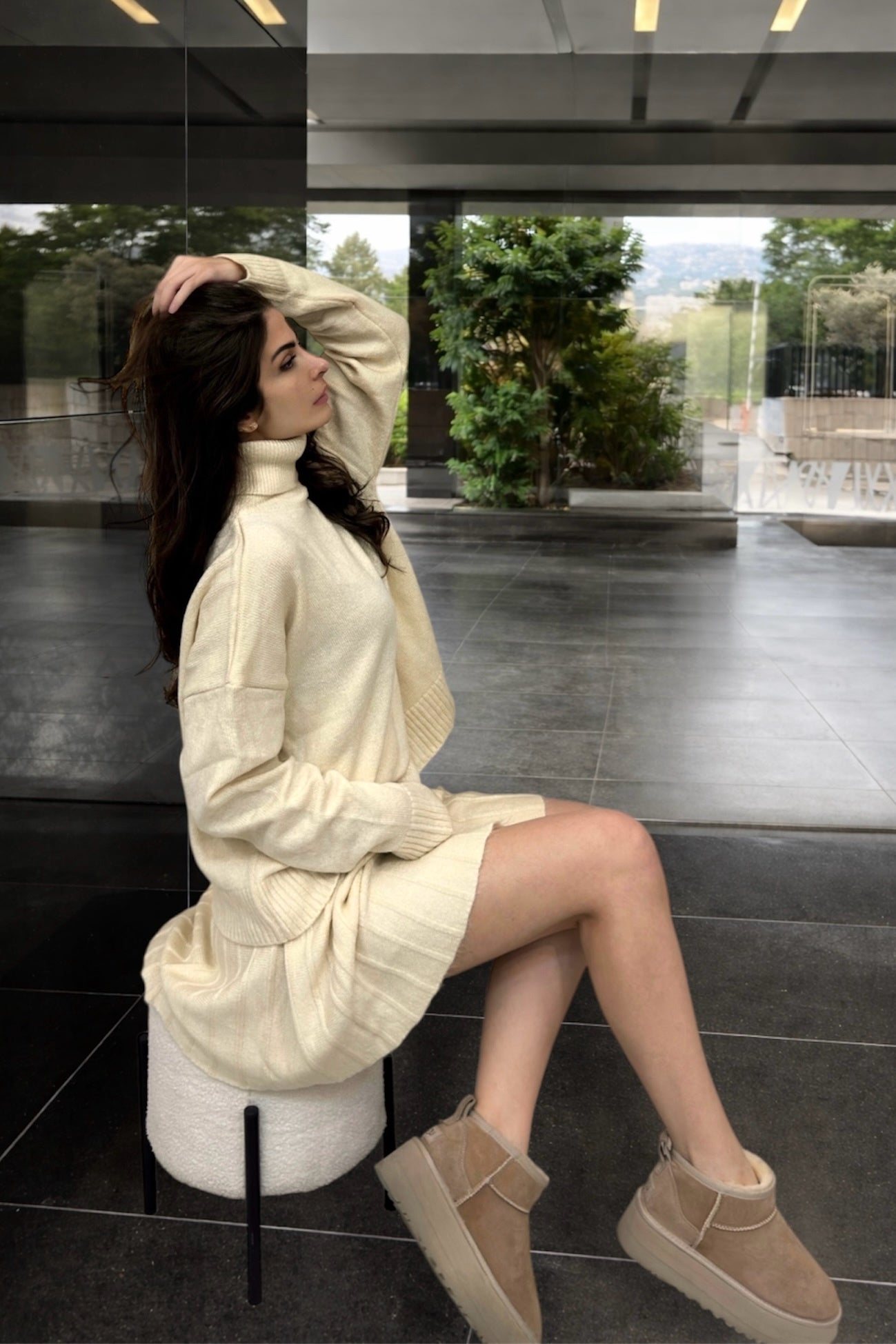 Woman in a cream outfit sitting on a stool in a modern indoor setting.