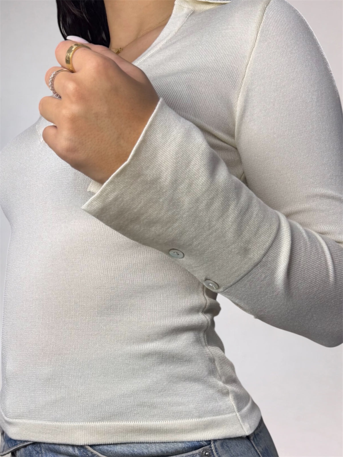 A close-up of a person wearing a light cream ribbed long-sleeve top with buttoned cuffs, paired with blue jeans. The person is wearing several rings on their fingers.