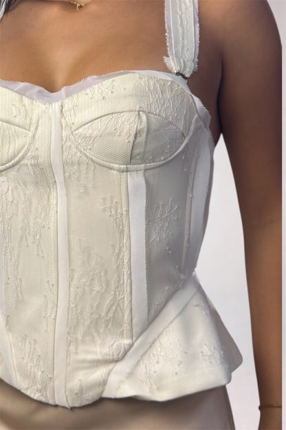 A close-up of a model wearing a structured white corset-style top with lace-like texture, satin trim, and a peplum hem against a plain light background.