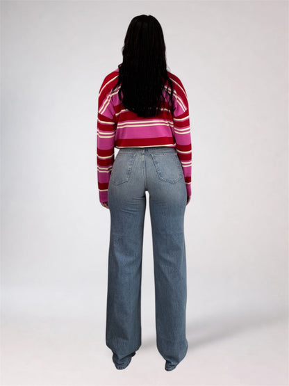 A woman viewed from behind wearing a red, pink, and white striped long-sleeve crop top and light blue wide-leg jeans, standing against a light gray studio background.