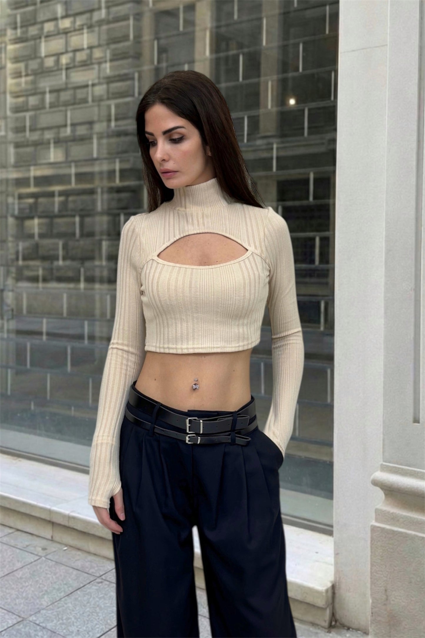 Woman wearing a beige ribbed top with cut-out design and black pants standing in front of a glass building.
