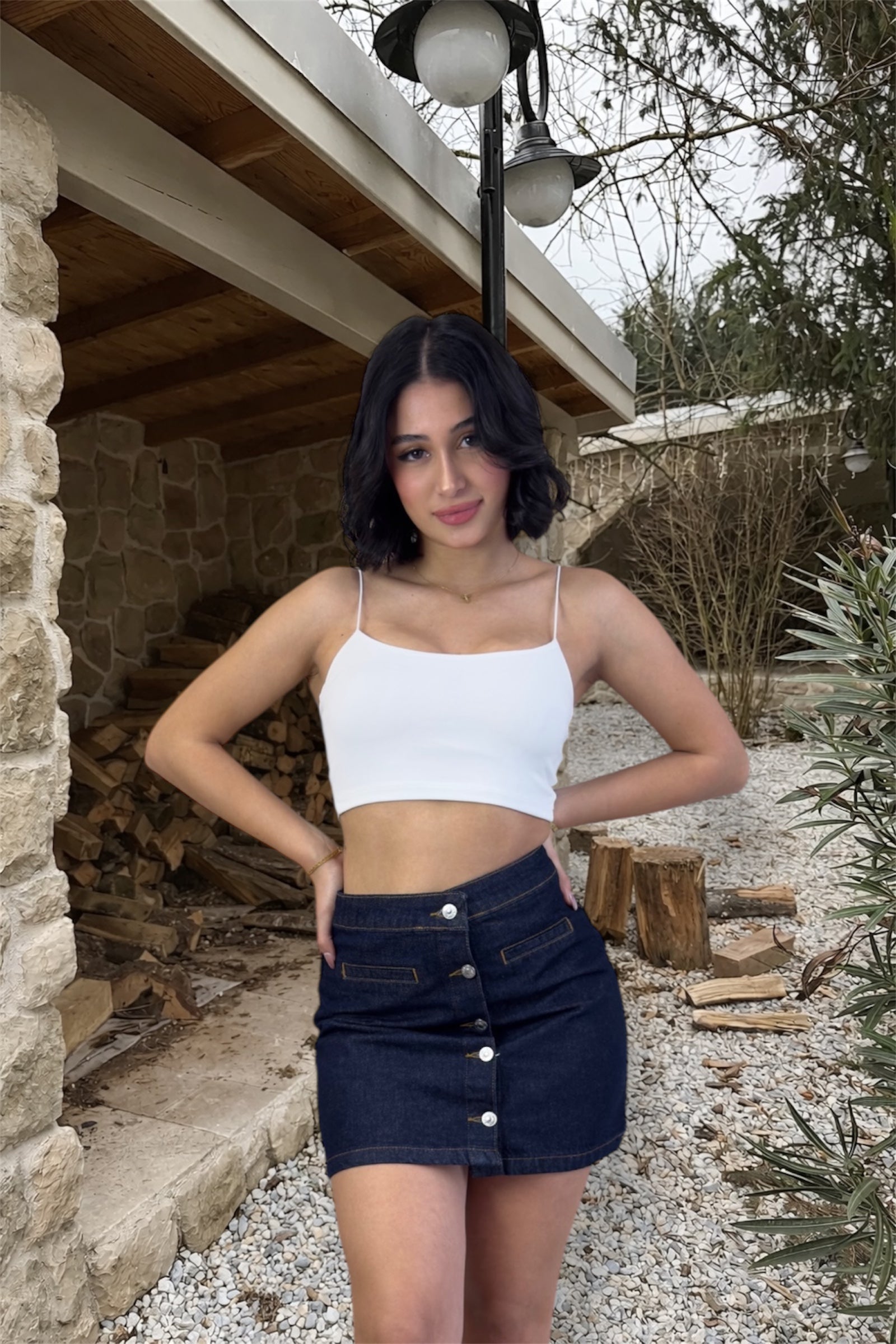 Woman in a white tank top and denim skirt standing outdoors near stone walls and wooden logs.