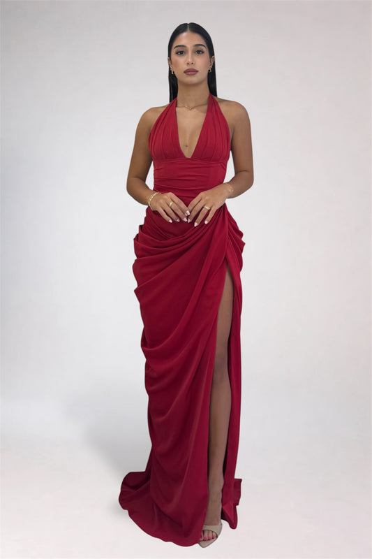A woman modeling a floor-length, deep red halter gown with a draped, gathered skirt and a high thigh slit, photographed against a plain light gray studio background.