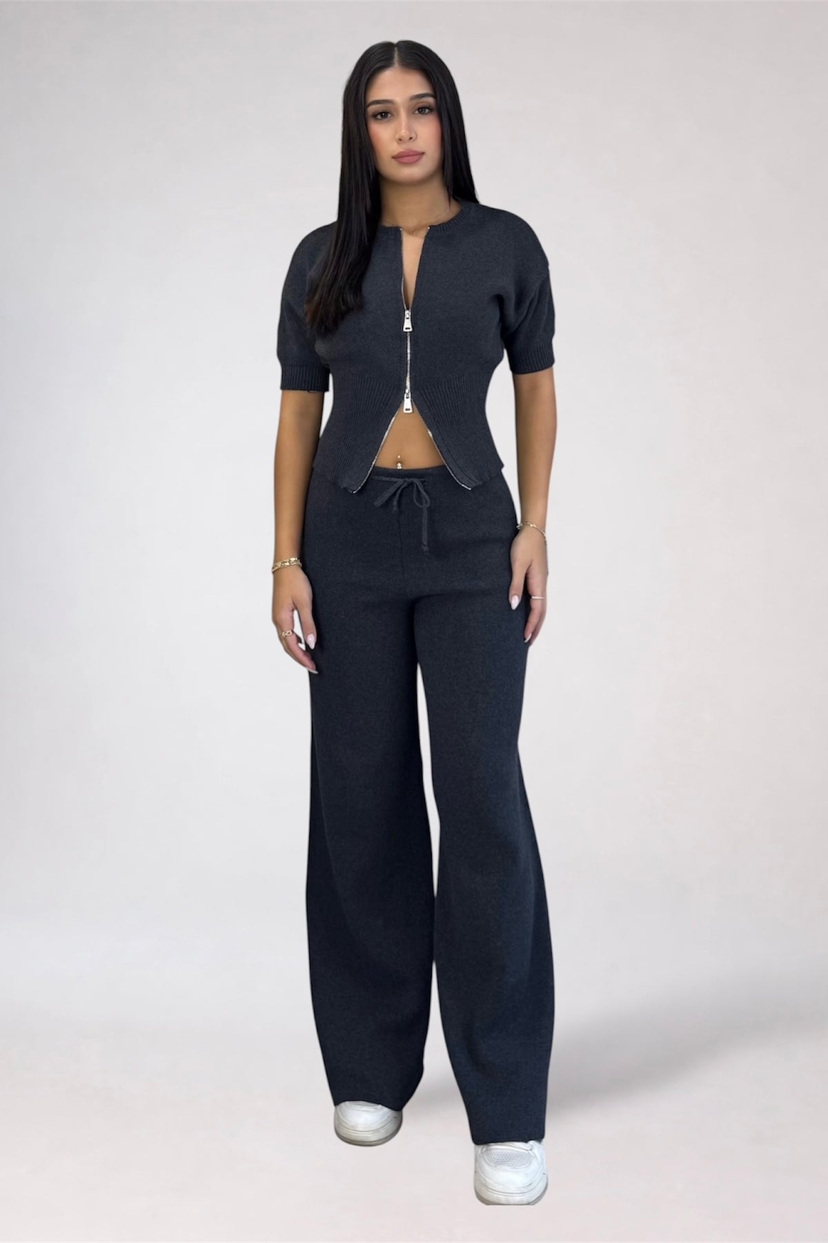 A woman modeling a dark navy blue two-piece knit set consisting of a short-sleeve cropped top with a front zipper and drawstring waist, paired with matching wide-leg pants, against a light gray studio background.