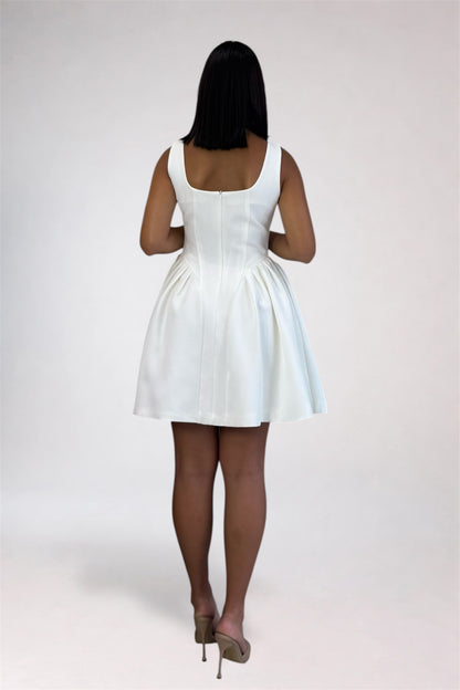 A woman viewed from behind wearing a white sleeveless mini dress with a structured back and gathered skirt, paired with beige high heels, against a plain light gray studio background.