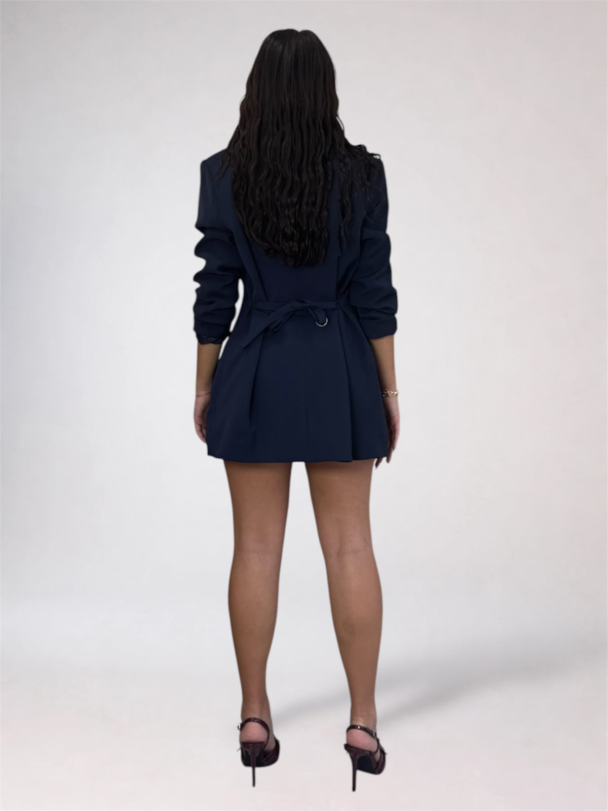 A woman viewed from behind wearing a dark navy blue belted blazer dress with long sleeves and black high heels, standing against a light gray studio background.