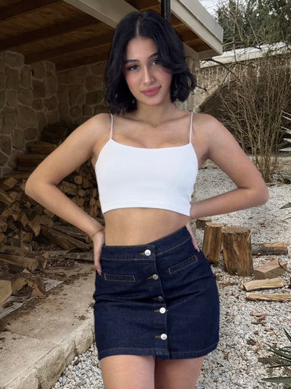 Woman wearing a white crop top and dark denim skirt standing outdoors with wooden logs and a stone wall in the background.