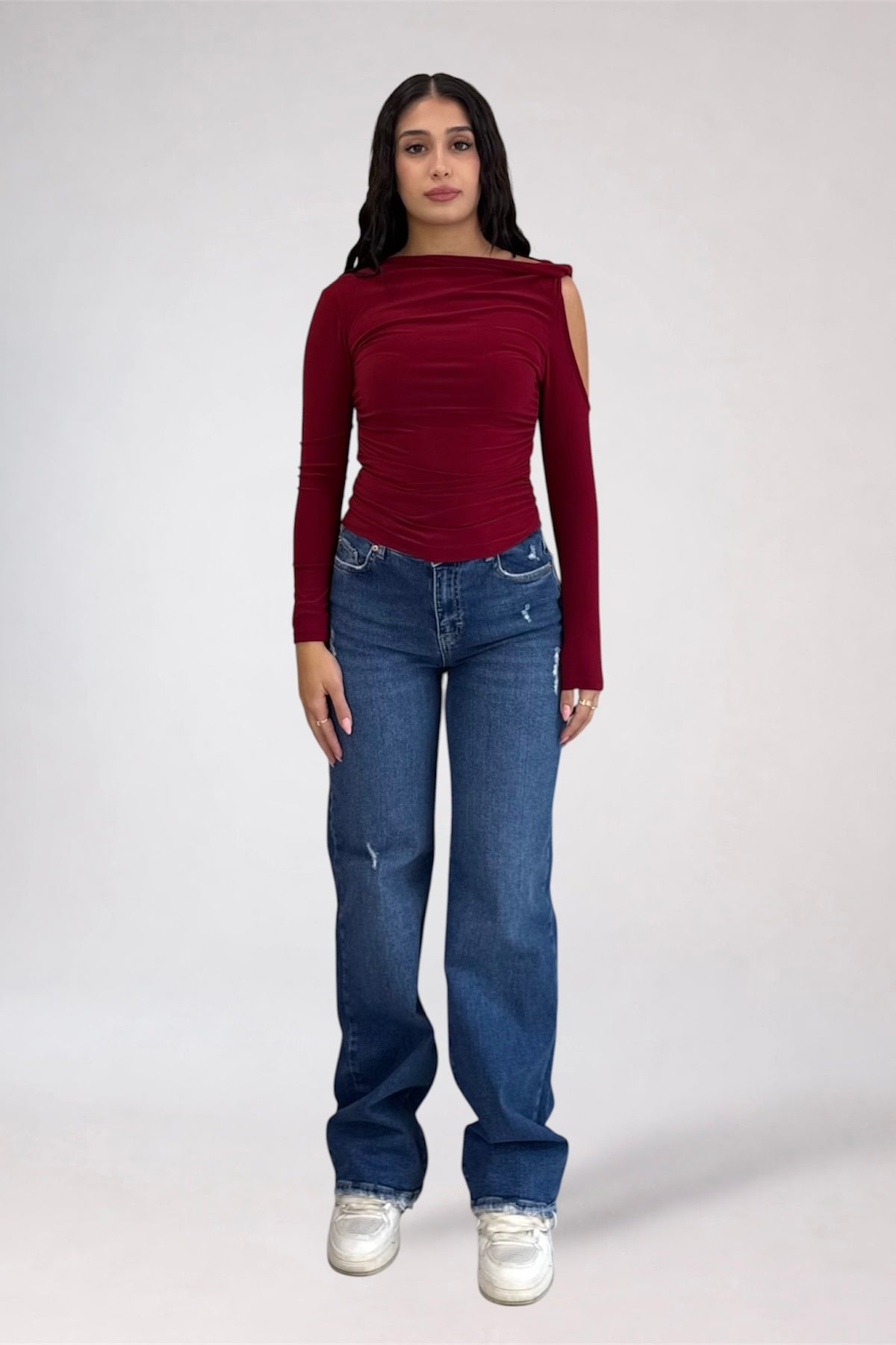 A woman modeling a dark red long-sleeve top with a draped neckline and cold-shoulder detail, paired with blue jeans and white sneakers, against a light gray studio background.