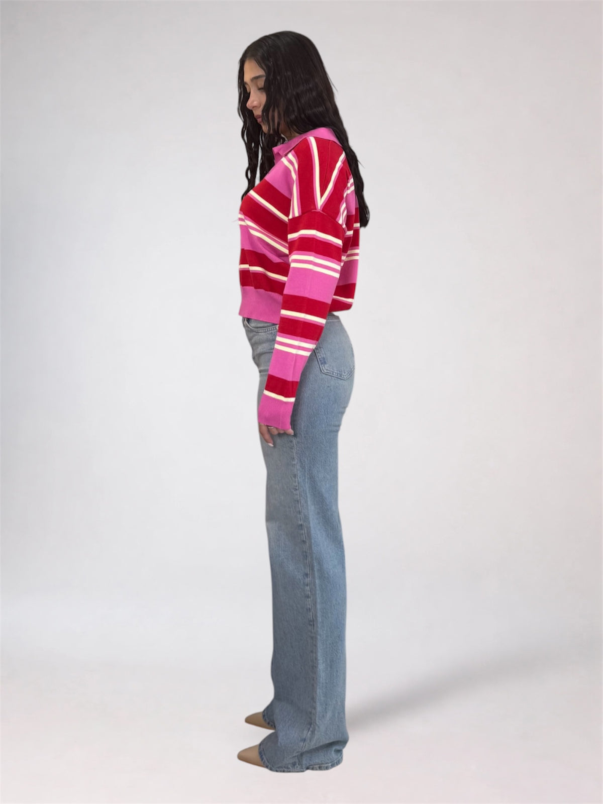 A woman stands in profile wearing a cropped knit sweater with bold horizontal stripes in red, pink, and white, paired with light blue flared jeans against a plain light gray studio background.