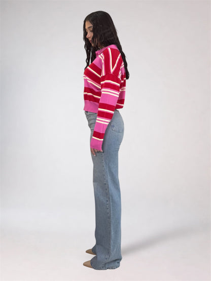 A woman stands in profile wearing a cropped knit sweater with bold horizontal stripes in red, pink, and white, paired with light blue flared jeans against a plain light gray studio background.