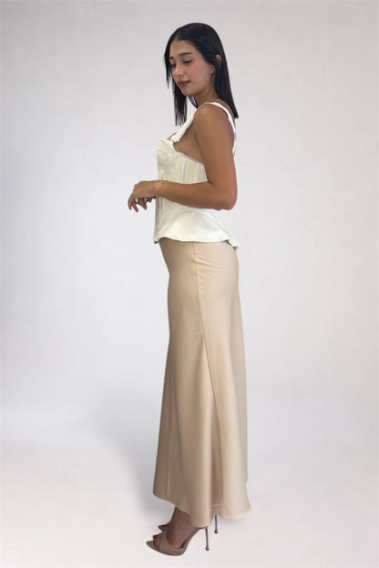 A woman modeling a two-piece outfit consisting of a cream sleeveless top and a beige maxi skirt, standing against a plain light gray studio background.