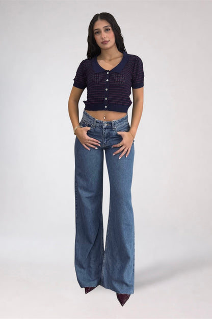 A woman modeling a dark navy blue short-sleeve cropped knit top with a button front and wide-leg blue jeans against a light gray studio background.