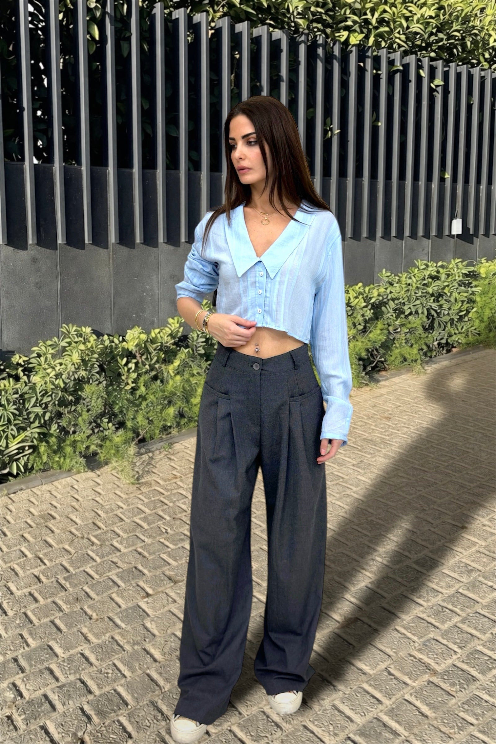 Woman wearing a light blue shirt and dark pants standing on a paved path with greenery in the background