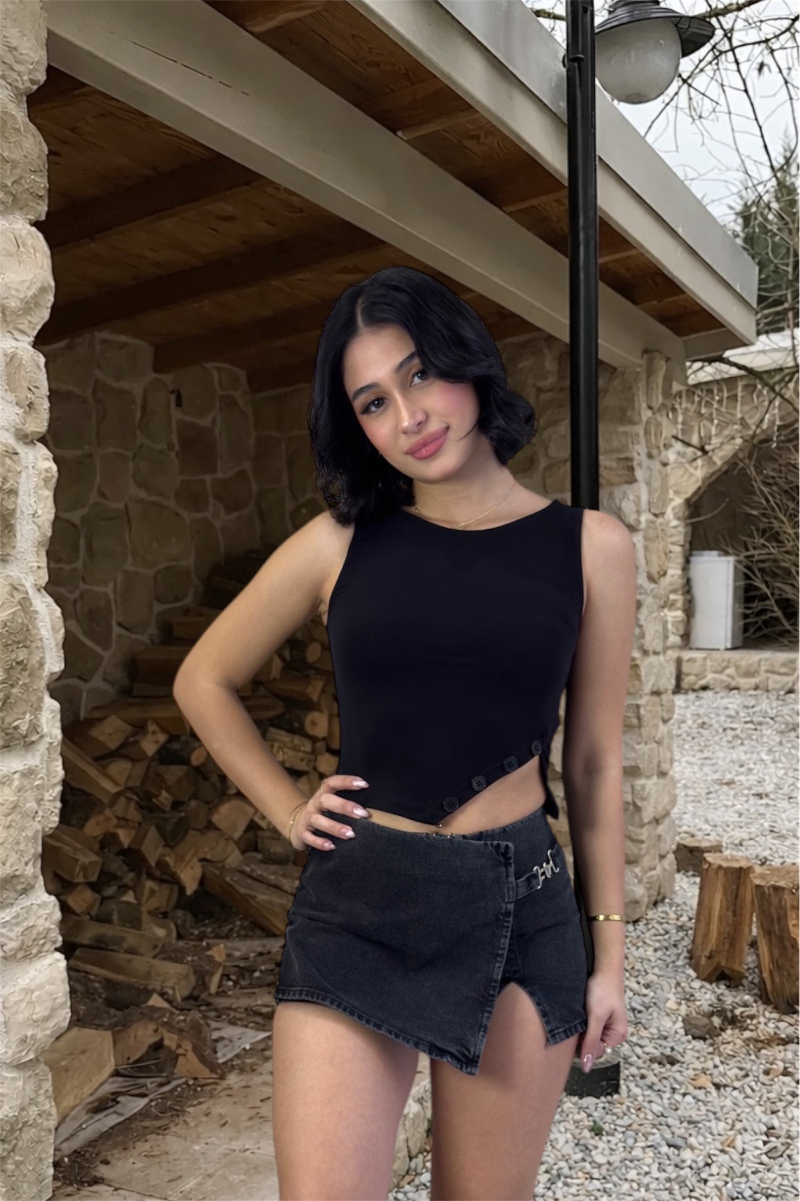 Woman in black sleeveless top and denim shorts standing outdoors near stone walls and wooden logs.