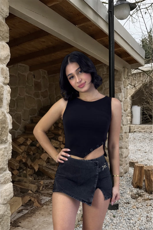 Woman in black sleeveless top and denim shorts standing outdoors near stone walls and wooden logs.