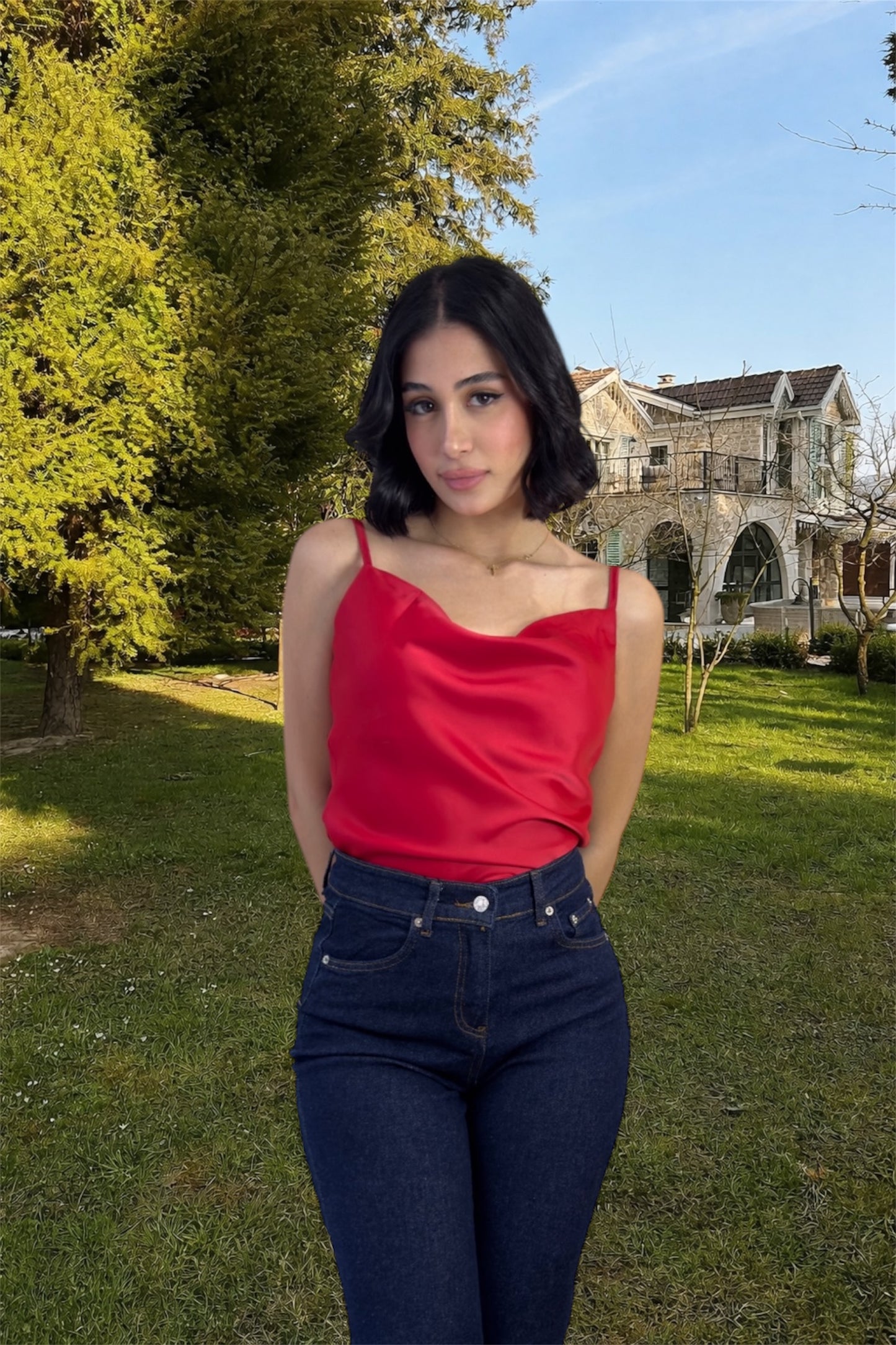 Woman in a red top and blue jeans standing outdoors with greenery and a building in the background