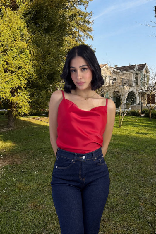 Woman in a red top and blue jeans standing outdoors with greenery and a building in the background