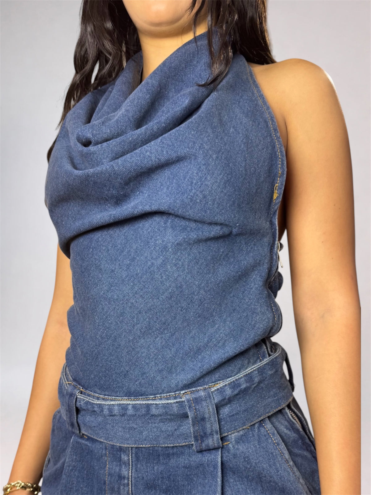 A close-up of a model wearing a blue denim halter top with a draped cowl neckline and matching high-waisted denim bottoms against a light gray background.