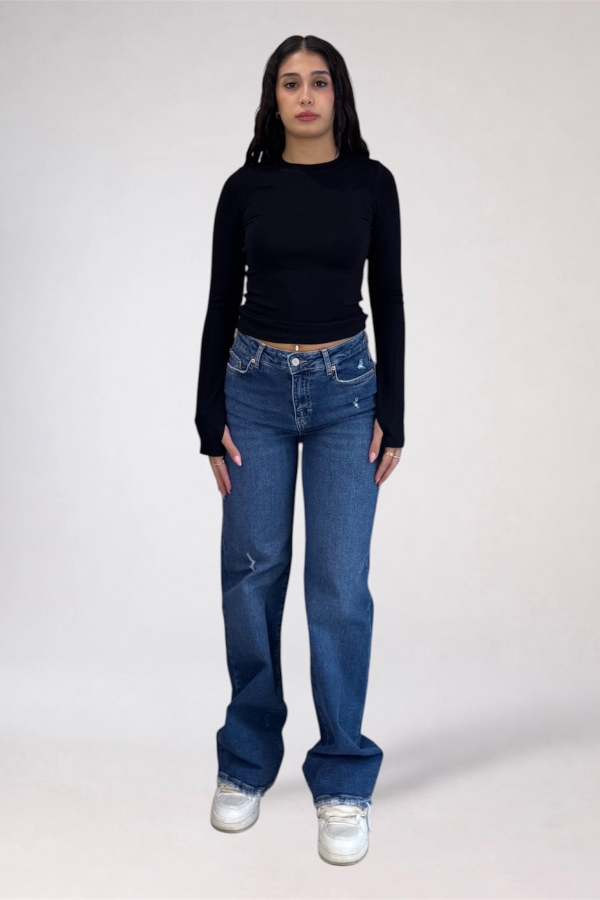 A woman modeling a black long-sleeve crop top and blue straight-leg jeans with white sneakers, standing against a light gray studio background.
