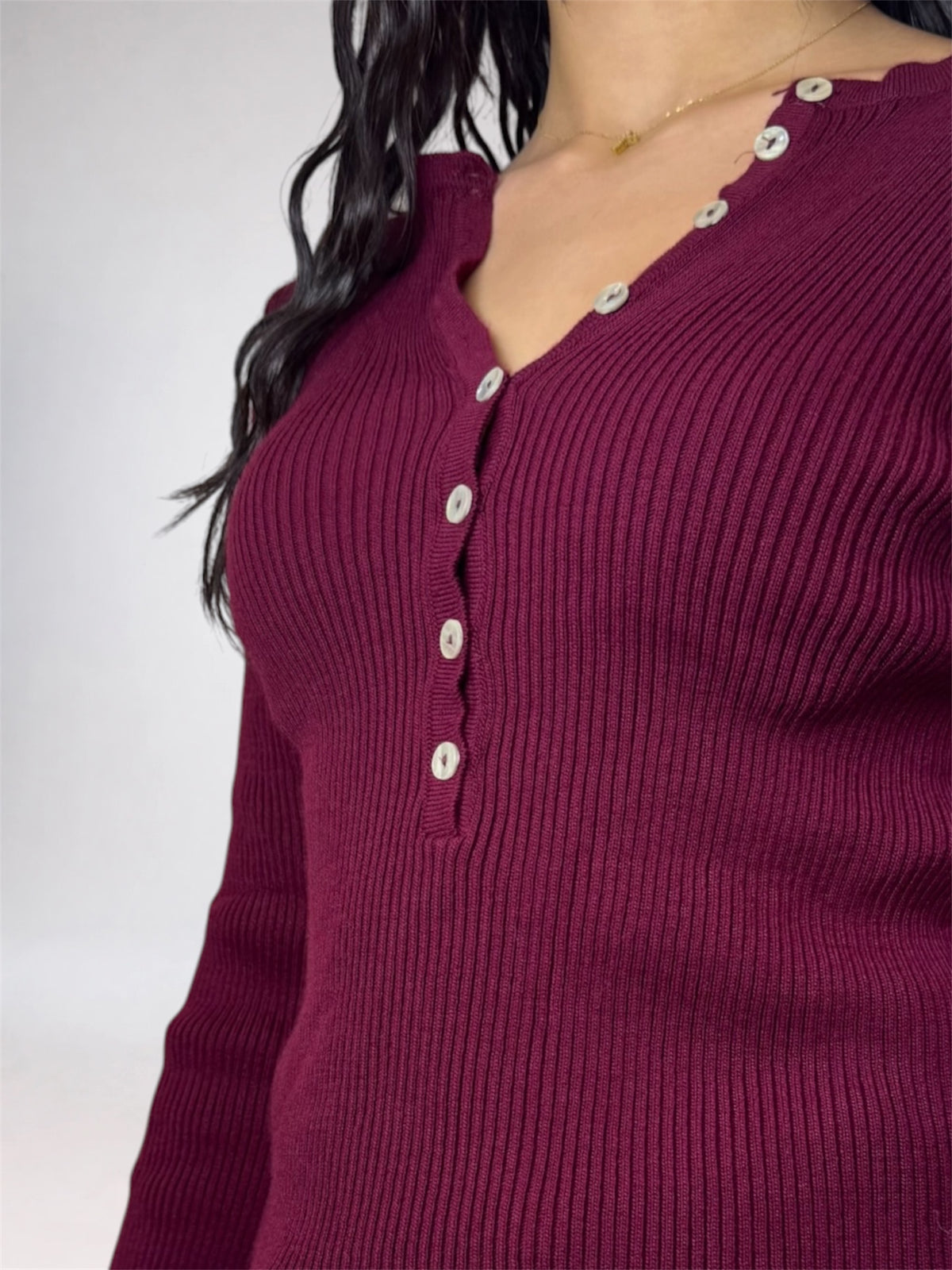 A close-up of a ribbed knit long-sleeve top in deep burgundy, featuring a split V-neckline with decorative silver-tone buttons down the front.