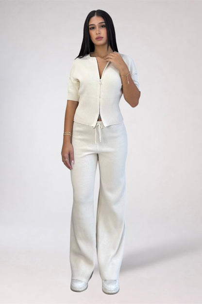 A woman modeling a matching off-white knit two-piece set consisting of a short-sleeve cropped zip-front top and wide-leg drawstring pants, photographed against a plain light gray studio background.