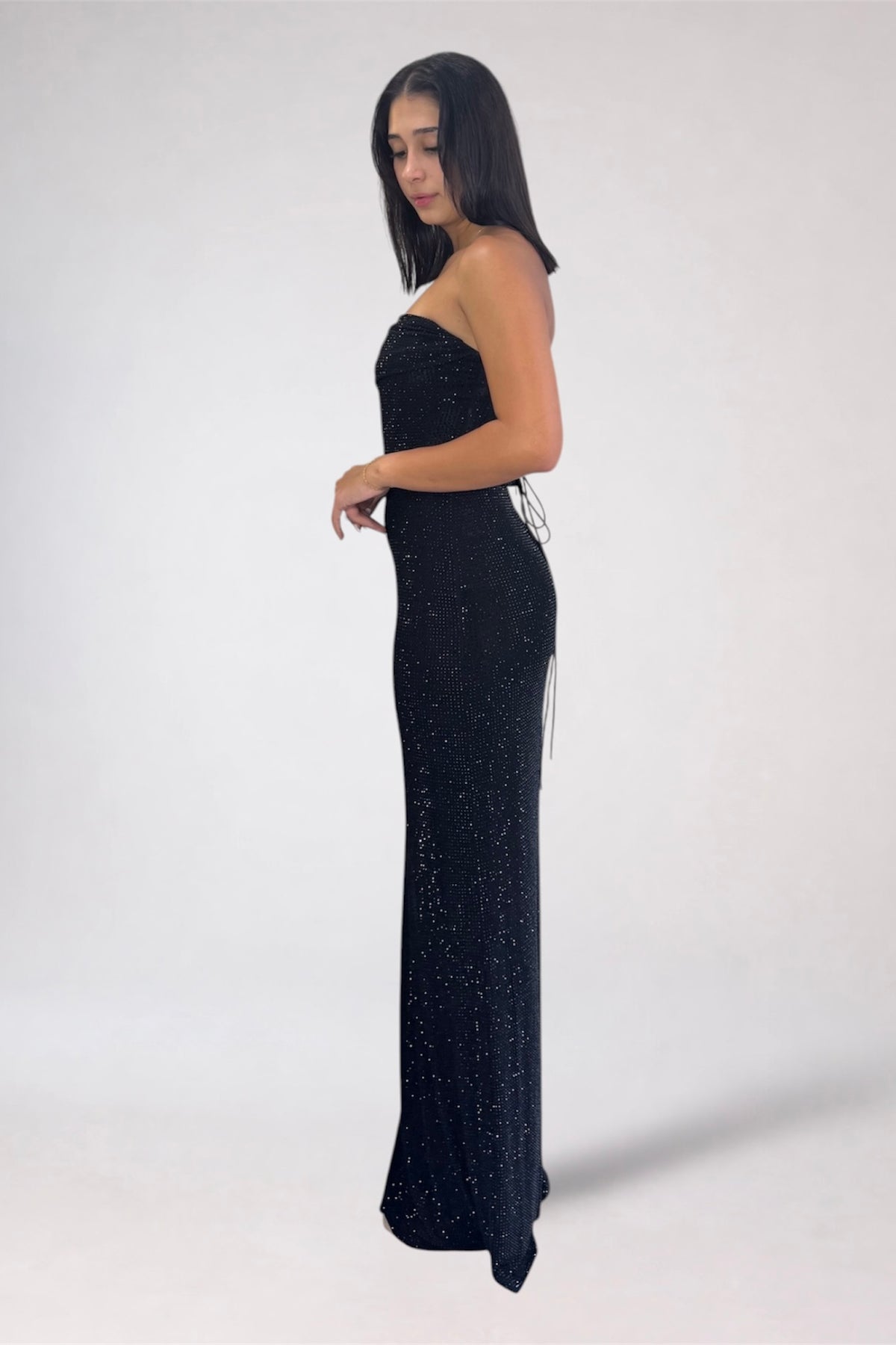 A woman modeling a strapless, floor-length black gown covered in shimmering sequins, shown in profile against a plain light gray studio background.