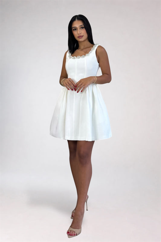 A woman modeling a sleeveless white mini dress with a pleated skirt and a beaded neckline, standing against a light gray studio background.