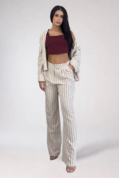 A woman modeling a matching beige and white pinstripe lounge set, consisting of a cropped long-sleeve top and wide-leg pants, styled with a dark red crop top and an open striped jacket against a plain light gray background.
