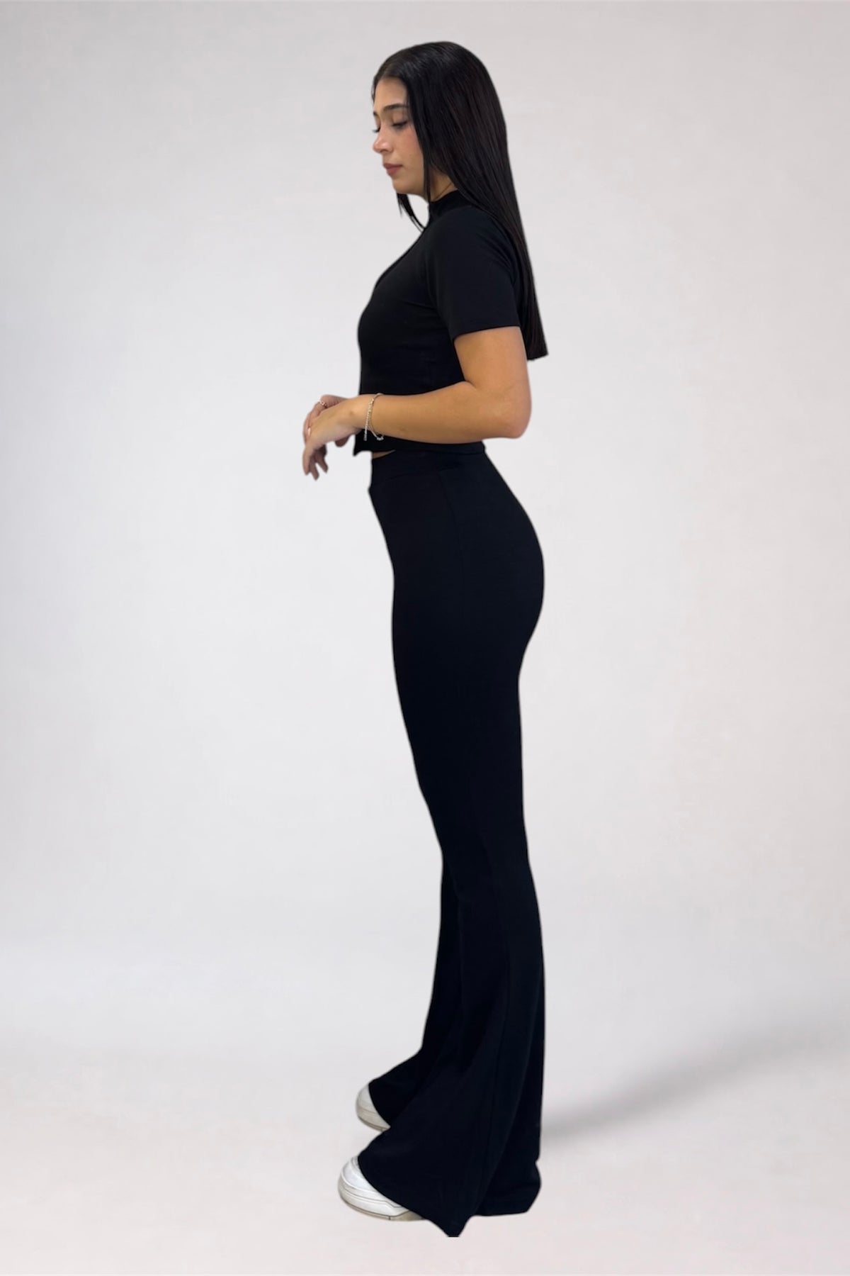 A woman modeling a black short-sleeve mock-neck jumpsuit with a fitted waist and flared legs, shown in profile against a light gray studio background.