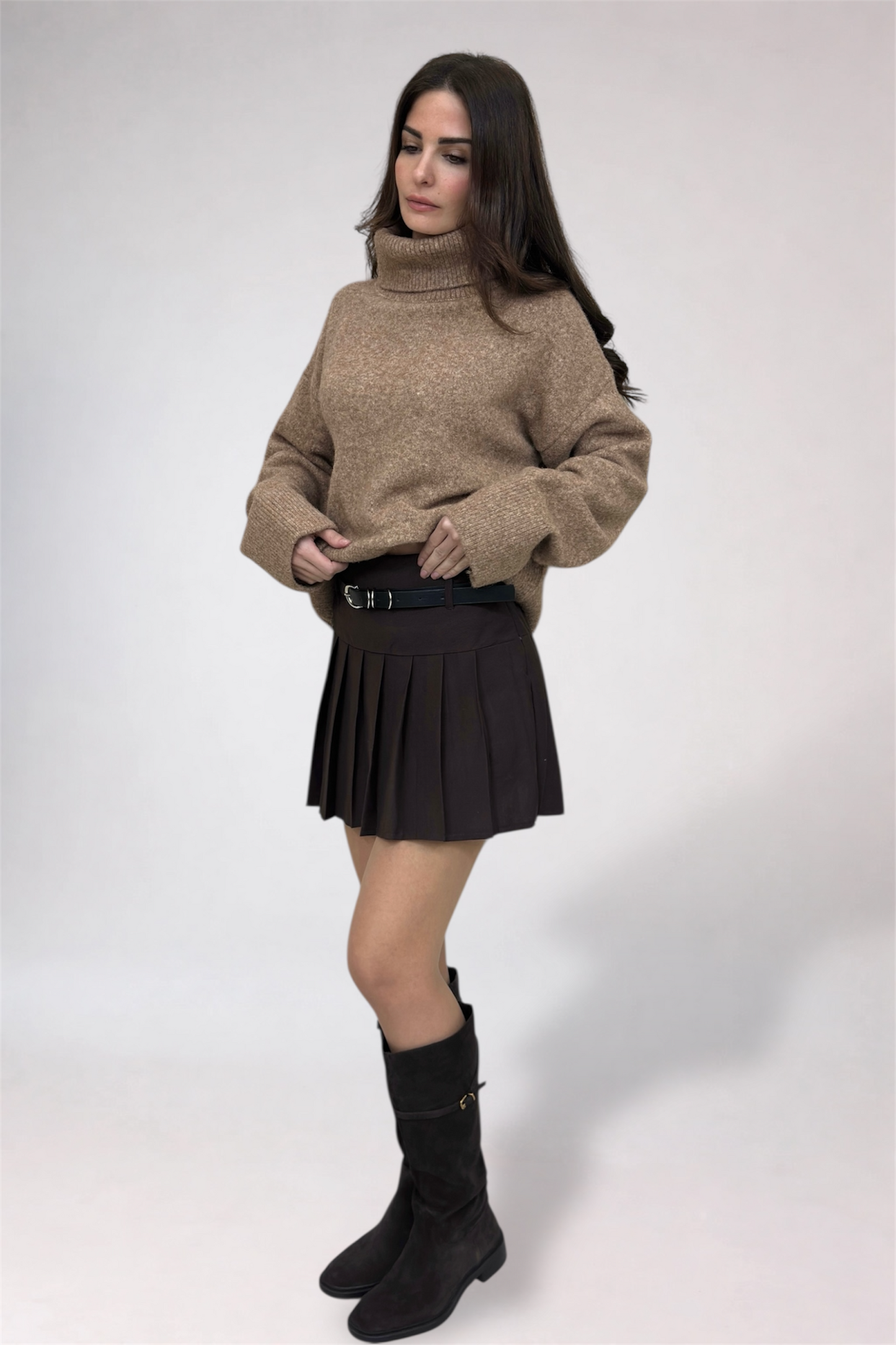 Woman wearing a beige sweater, black pleated skirt, and black boots on a white background
