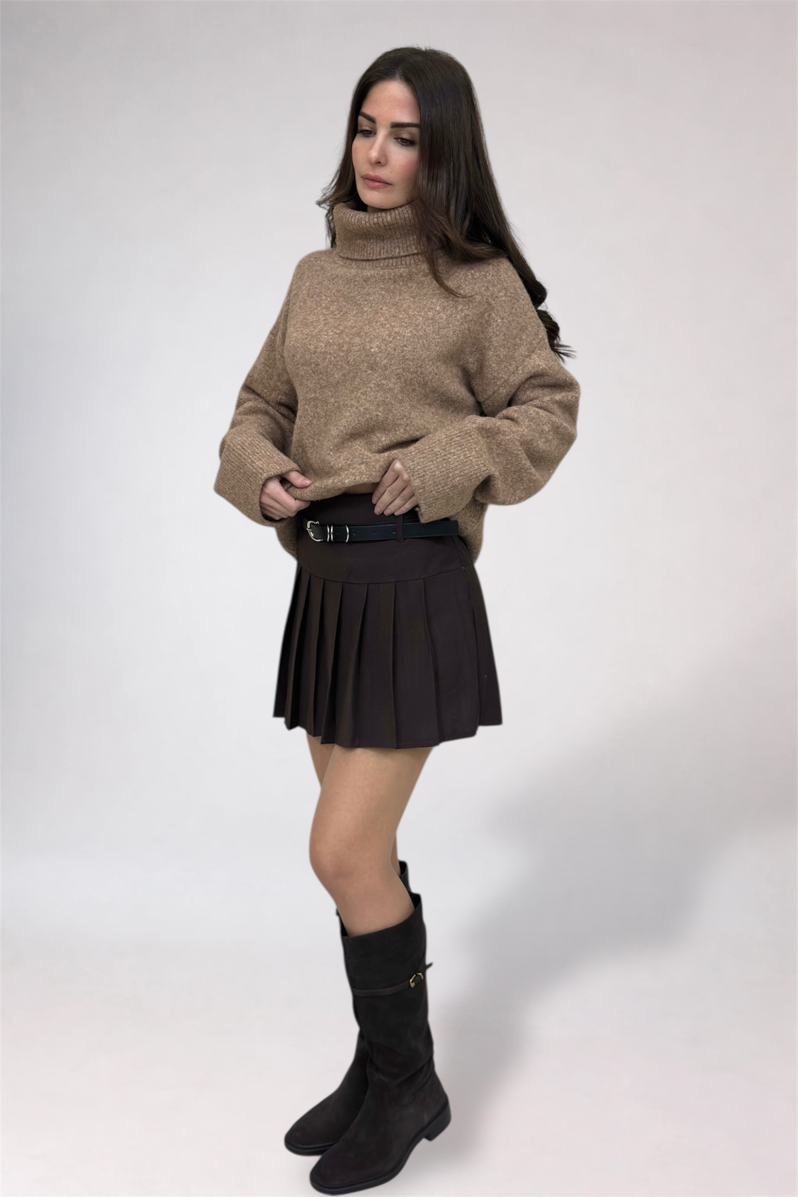 Woman wearing a beige sweater, black pleated skirt, and black boots on a white background