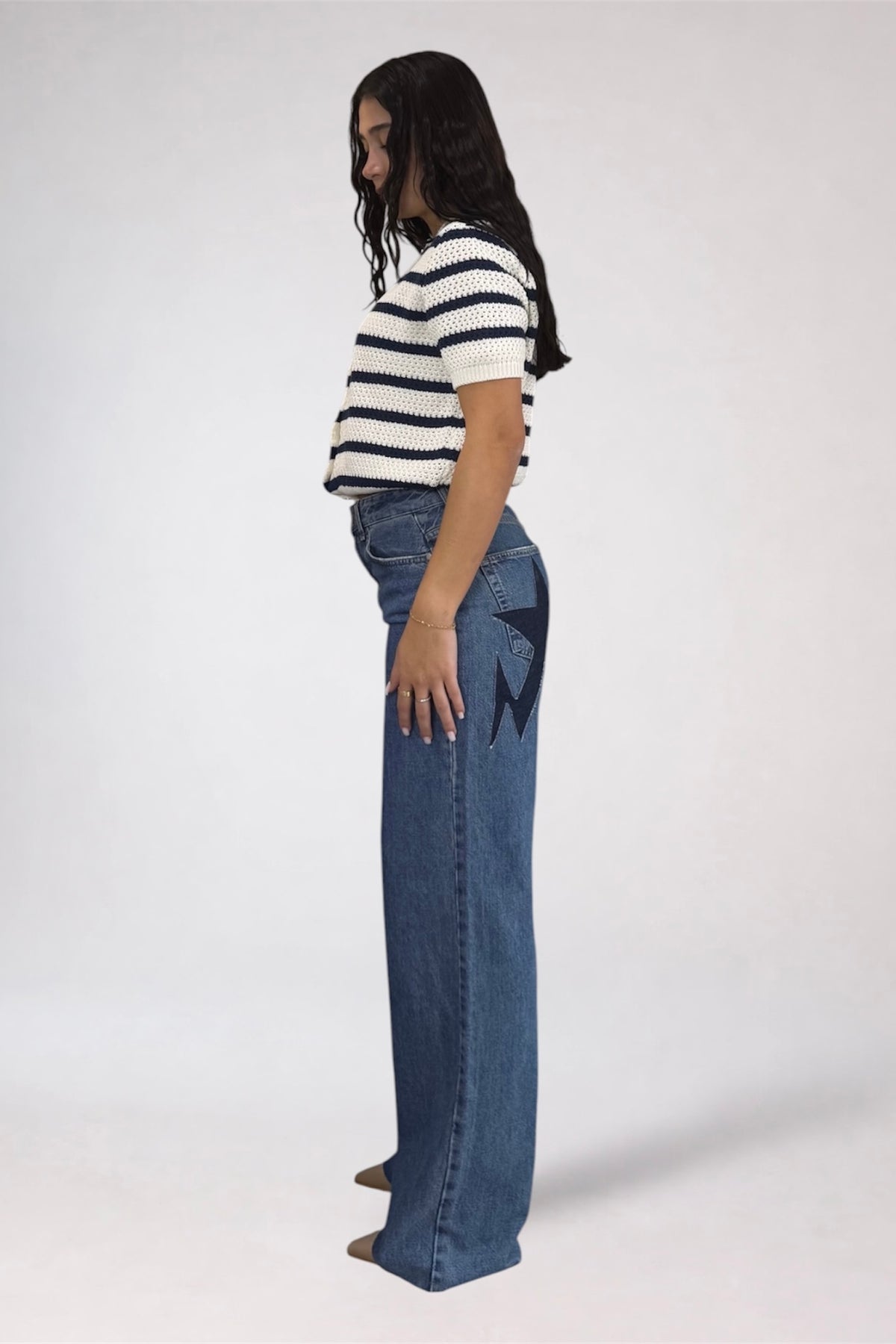 A woman shown from the side profile wearing a black and white striped short-sleeve crop top and high-waisted, wide-leg blue denim jeans with a black graphic print on the back pocket, set against a plain light gray studio background.