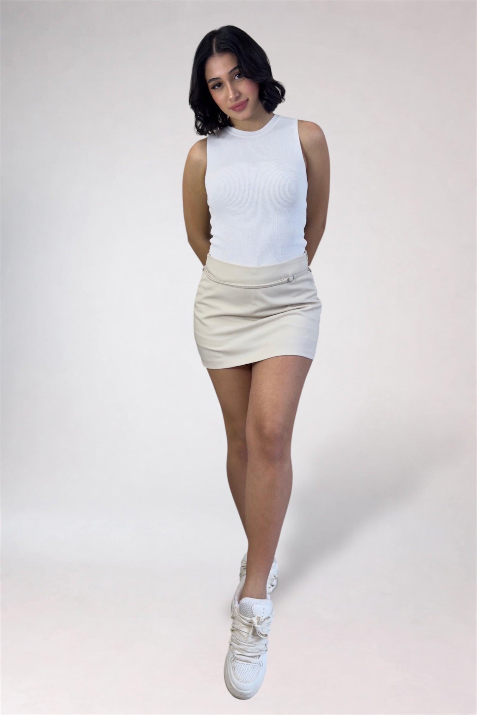 Person wearing a white sleeveless top and beige skirt on a plain background