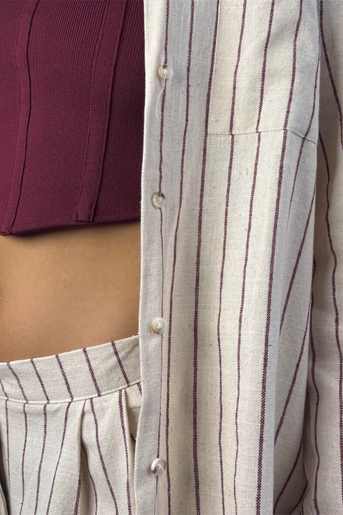 Close-up of a person wearing a light beige linen button-front shirt with thin dark vertical pinstripes, paired with matching striped bottoms and a burgundy ribbed crop top.