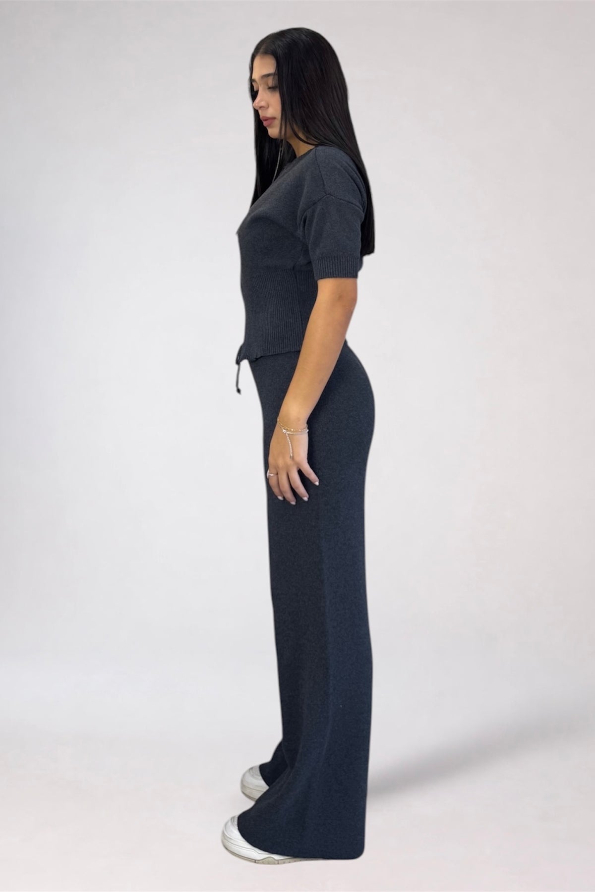 A woman modeling a dark charcoal gray knit two-piece set consisting of a short-sleeve crop top and matching wide-leg pants, shown in profile against a light gray studio background.