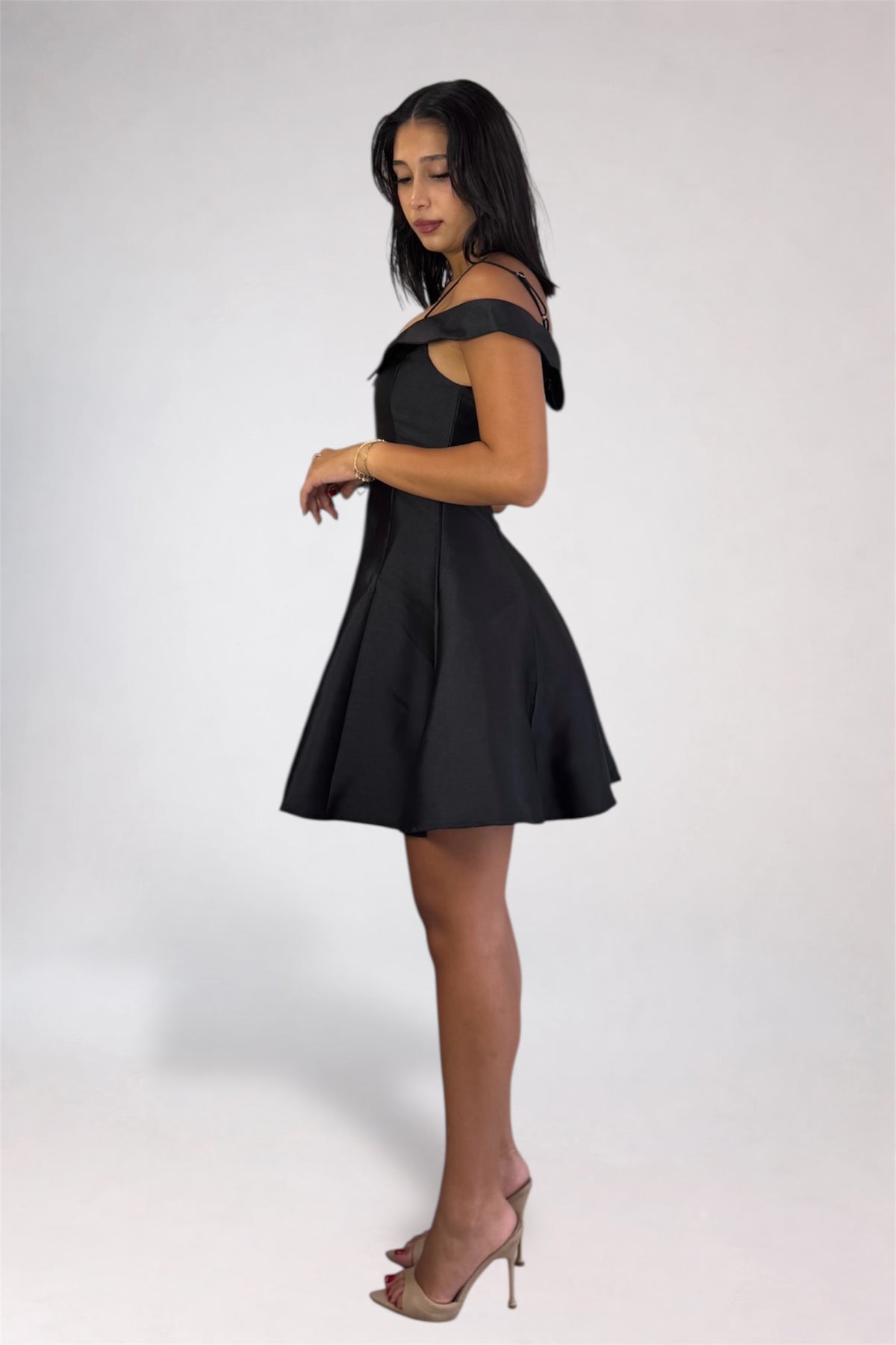 A woman modeling a short black off-the-shoulder dress with a flared skirt, paired with nude high-heeled sandals, against a plain light gray studio background.