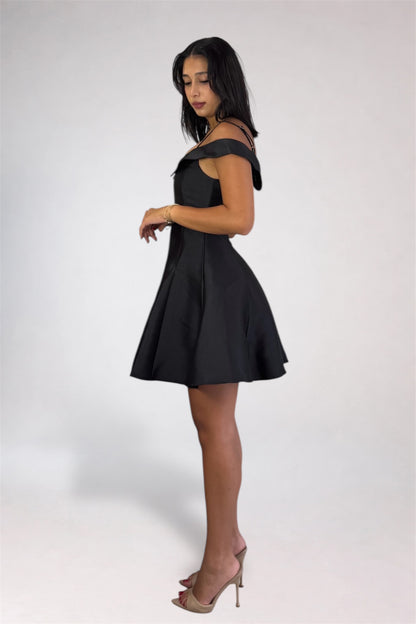 A woman modeling a short black off-the-shoulder dress with a flared skirt, paired with nude high-heeled sandals, against a plain light gray studio background.